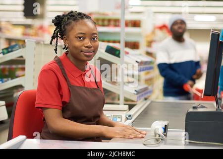 Happy young black woman in red shirt and brown apron sitting by counter with scanner and computer screen showing price of goods Stock Photo