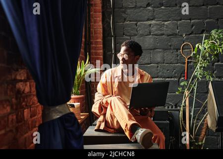 Young serious African American man with laptop sitting by windowsill and looking through window while concentrating on work Stock Photo