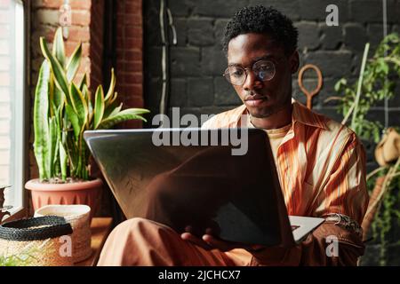 Young serious black man in casualwear and eyeglasses using laptop while sitting by window in living room of modern loft apartment Stock Photo