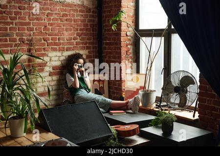 Cute girl in casualwear sitting by windowsill with air flow fan, domestic plant in flowerpot and picture in frame and taking photo of living room Stock Photo