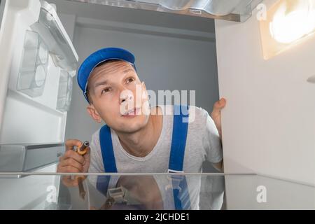 Male technician in overalls repairs a broken refrigerator with a screwdriver. Refrigerator repair concept. Photo from inside the refrigerator Stock Photo