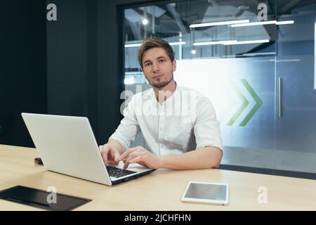 Portrait of young businessman with beard, man working in modern office with laptop smiling and looking at camera Stock Photo