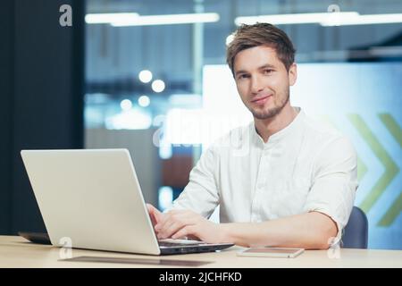 Portrait of young businessman with beard, man working in modern office with laptop smiling and looking at camera Stock Photo
