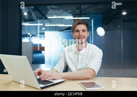 Portrait of young businessman with beard, man working in modern office with laptop smiling and looking at camera Stock Photo