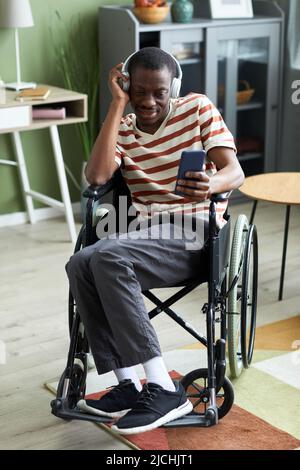 Full length portrait of smiling African American man with disability listening to music and using phone in modern home Stock Photo