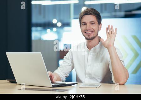 Portrait of young office worker, man looking at camera and smiling waving hand greeting gesture, working on laptop businessman Stock Photo