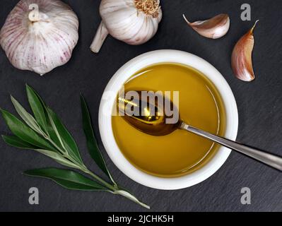 top view of a bowl of virgin olive oil on slate plate with garlic and olive tree branch Stock Photo