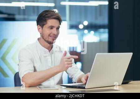 A man in the office works on a laptop, talks on a video call, looks at the screen and smiles at the interlocutor Stock Photo