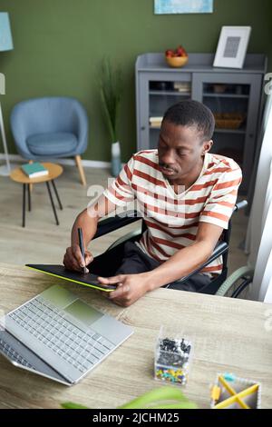 Vertical portrait of African American man in wheelchair using pen tablet while working with computer at home Stock Photo