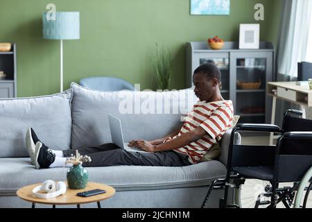 Side view portrait of African American man with disability using laptop at home while lying on couch, wheelchair in foreground Stock Photo