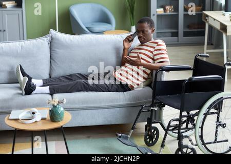 Side view portrait of African American man with disability speaking on phone at home while lying on couch, wheelchair in foreground Stock Photo