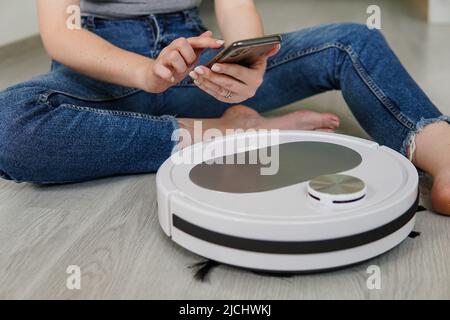 Woman housewife using mobile application to control robotic vacuum cleaner for start cleaning. Smart home concept. Stock Photo