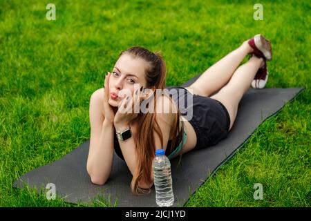 Cute caucasian woman wearing sportive clothes on city park, outdoors lying on yoga mat and looking away with cute expression. Outdoor sport and health Stock Photo