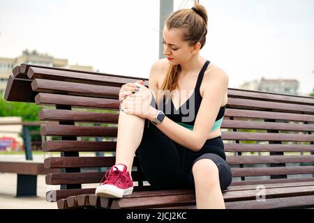 Young brunette woman wearing sportive clothes on city park, outdoors cramps in leg and feel pain, touching her knee ankle painful injury outside after Stock Photo