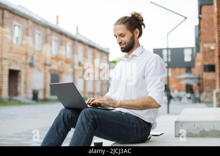 Stylish business guy using modern laptop for remote work while sitting on stairs near office building. Smiling male freelancer enjoying favorite work during break. Stock Photo