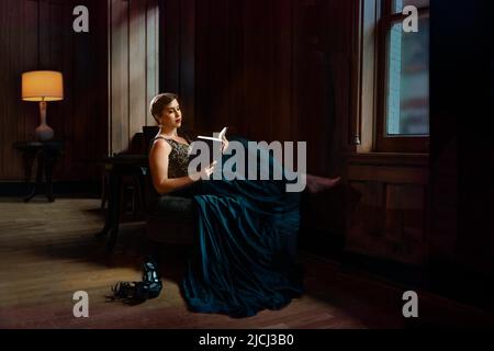 A young woman slips out of a party to find a quiet corner, reading a book. She is an introvert, escaping the crowds and noise in this peaceful room. Stock Photo