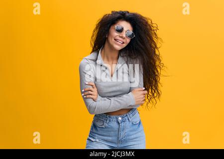 GOOD OFFER. Happy enjoyed pretty Latin female with afro in sunglasses hugging herself, laughing, stay isolated over yellow background. Copy space Stock Photo