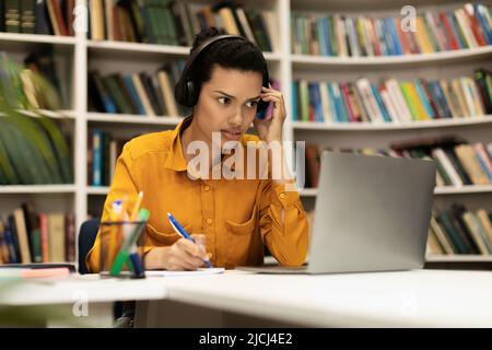 Focused female student looking at laptop, learning online and taking notes, sitting at desk in library Stock Photo