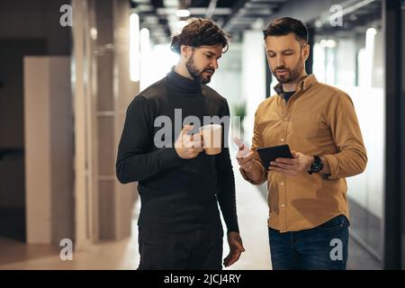 Male coworkers discussing business startup project, using digital tablet while walking in office hall on coffee break Stock Photo