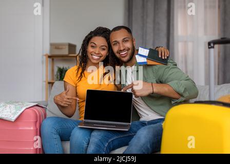 Travel website advertisement. Happy black couple showing laptop blank screen, sitting with packed suitcases, mockup Stock Photo