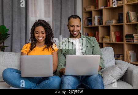 Young african american guy and his girlfriend using modern laptops and smiling while sitting on couch at home Stock Photo