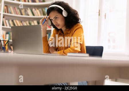 Focused mixed race lady studying online with laptop and wireless headset, sitting in library, copy space Stock Photo