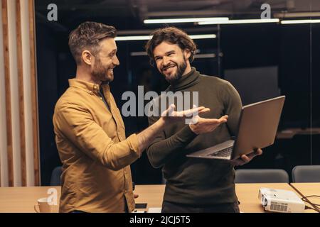 Portrait of two cheerful male colleagues working on startup project together, discussing ideas, using laptop at office Stock Photo