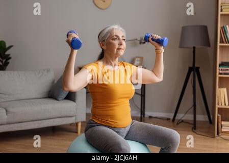 Stay home, do sports. Fit senior woman doing exercises on fitness ball and using dumbbells, working out at home Stock Photo
