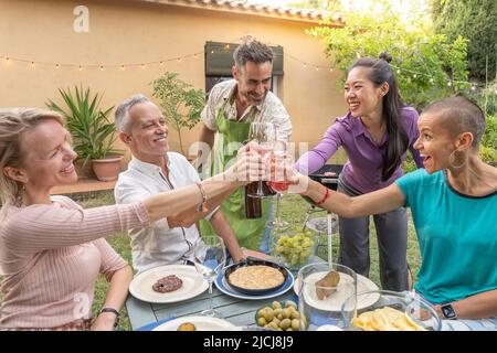 Multiracial middle aged friends eating, celebrating and toasting together in summer barbecue party. Stock Photo