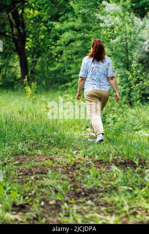 Back view of young plump buxom woman with long curly red hair walking on path in park forest among green trees. Summer. Stock Photo