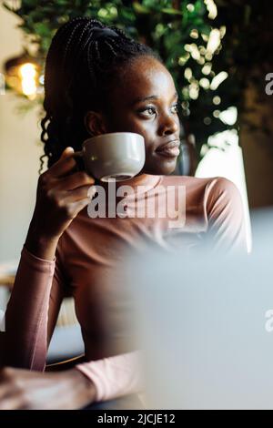 Vertical photo calm, dreaming african afro american young woman with dreadlocks, drinking coffee in cafe. Coffee break Stock Photo