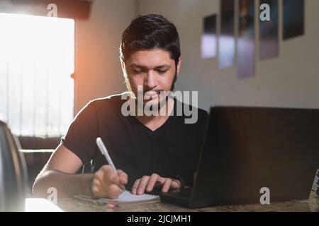 Concentrated young student writes in a notebook while learning by watching a video on the computer in his living room. An independent thinking man wri Stock Photo