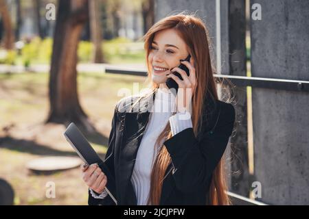 Modern red-haired businesswoman talking on phone sitting on bench in spring park outdoors, holding laptop. People urban lifestyle concep. Female using Stock Photo