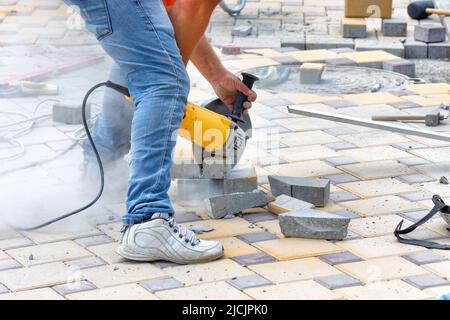 A worker using an electric cutter and a cutting disc cuts paving slabs during the laying of the sidewalk in the daytime. Copy space. Stock Photo