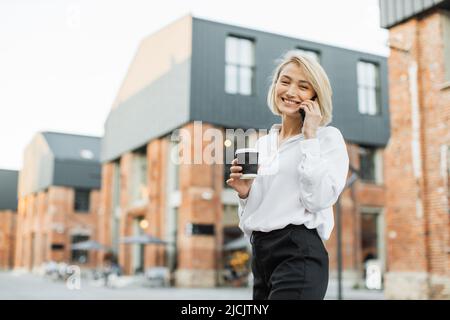 Portrait of beautiful blond woman having cup of fresh take away beverage talking to partner using smartphone walking on city street looking outside camera. Stock Photo