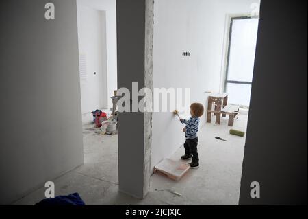 Cute male child painting white wall between two rooms under renovation. Adorable baby boy using paint brush while renovating apartment. Concept of home renovation, restoration and childhood. Stock Photo
