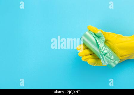 A hand in a yellow glove holds a plastic disposable trash bag on a blue background. place with copy space. Stock Photo