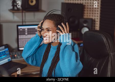 Cheerful funny African American woman presenter cutely smiles while putting on headphones, ready to speak with listeners Stock Photo