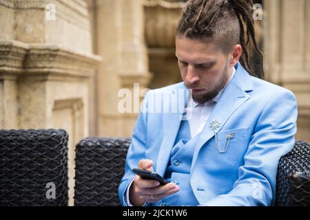 A young hipster man with dreadlocks in a blue suit sits in a street restaurant looking at a smartphone, sad and upset Stock Photo