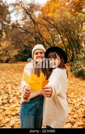 Two long-haired brown-haired women happily indulge, holding bright fallen leaves in park in clearing strewn with foliage. Golden autumn. Holidays Stock Photo