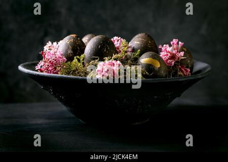 Black Easter concept. Bio colored black eggs with golden spots laying on wild moss with small pink flowers in black ceramic bowl on dark wooden backgr Stock Photo