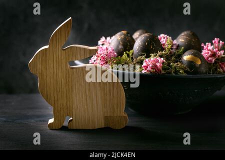 Black Easter concept. Bio colored black eggs with golden spots laying on wild moss with small pink flowers in black ceramic bowl, wooden rabbit bunny Stock Photo