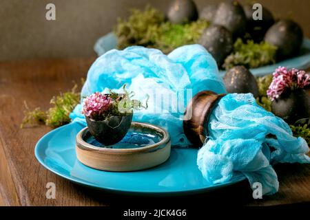 Easter table setting with empty turquoise ceramic plates, decorated by forest moss, black colored easter eggs and pink spring flowers in shell on wood Stock Photo