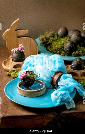 Easter table setting with empty turquoise ceramic plates, decorated by forest moss, wooden bunny black colored easter eggs and pink spring flowers on Stock Photo
