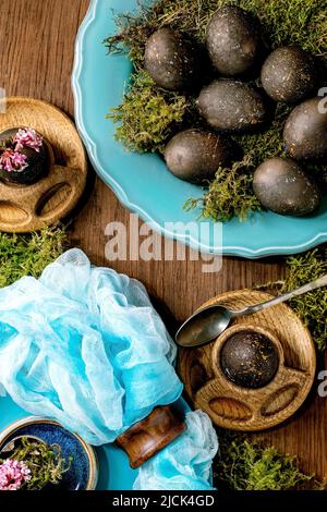 Easter table setting with empty turquoise ceramic plates and egg cups, decorated by forest moss, black colored easter eggs and pink spring flowers on Stock Photo