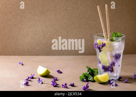 Transparent glass of lemonade soda cocktail with mint, violets flower ice and lime slices, paper straws, standing on brown table. Cold summer drink Stock Photo