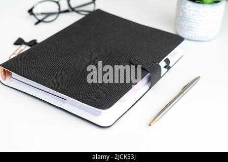 Close up picture of black notepad on white table with glasses, pen and flower aside. Mockup blank space copy space black notebook planner journal Stock Photo