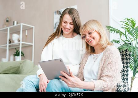 Senior woman and daughter playing with tablet pc. Smiling older woman and her granddaughter sitting home using gadget digital device for online Stock Photo