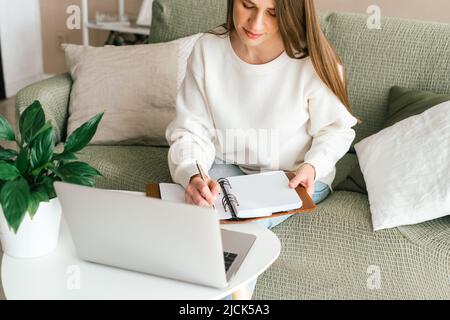 Thinking Freelance caucasian woman writing in notebook, typing at laptop and working from home office with plants. Happy girl with notepad on Stock Photo