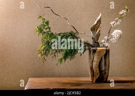 Beautiful spring ikebana. Floral arrangement with spring blooming white flowers, thuja branch and piece of wood in brown ceramic vase, standing on woo Stock Photo